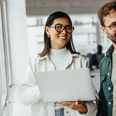 Business woman holding a laptop and standing with her colleague in an office. Two happy business people working together in a creative workplace.